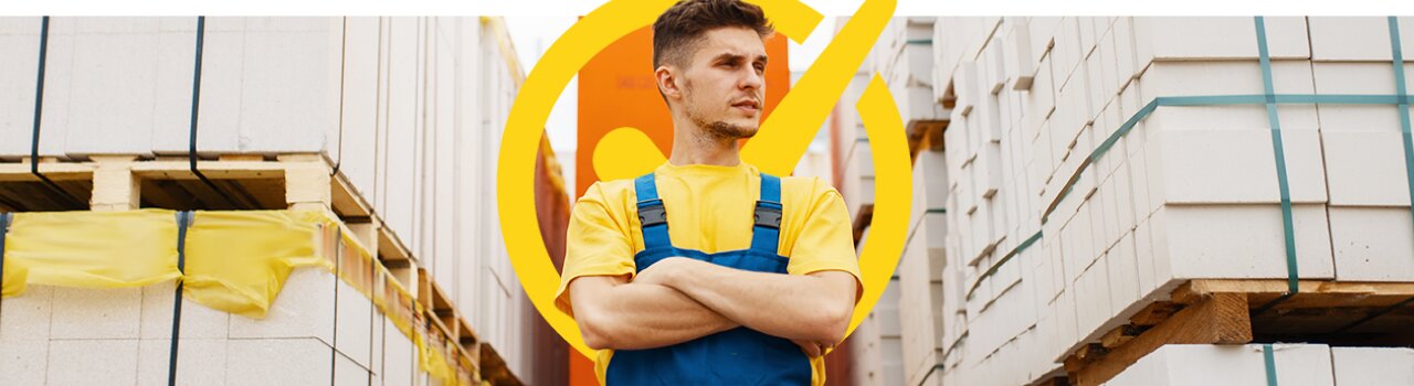 Man in yellow shirt and blue overalls from EWI Pro standing with crossed arms between stacks of aerated concrete blocks on pallets, with a yellow check mark symbol behind him